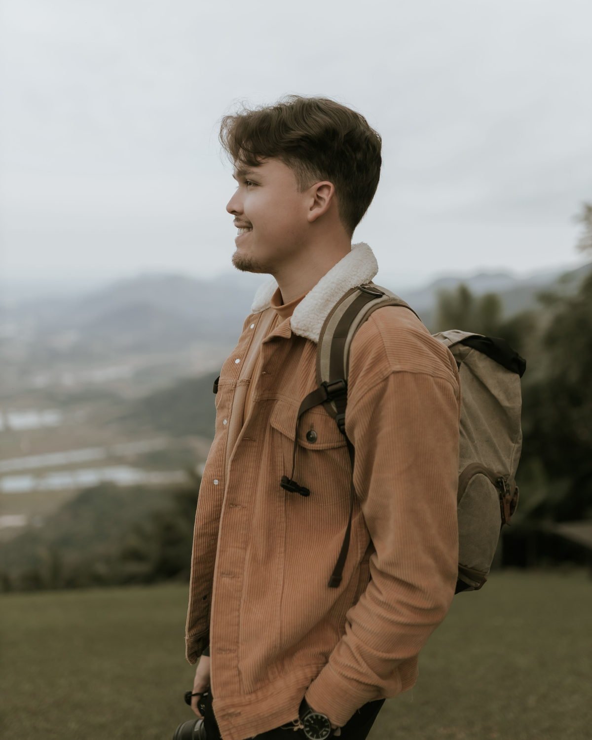 Adi outdoors with a camera slung over one shoulder, mountains behind him