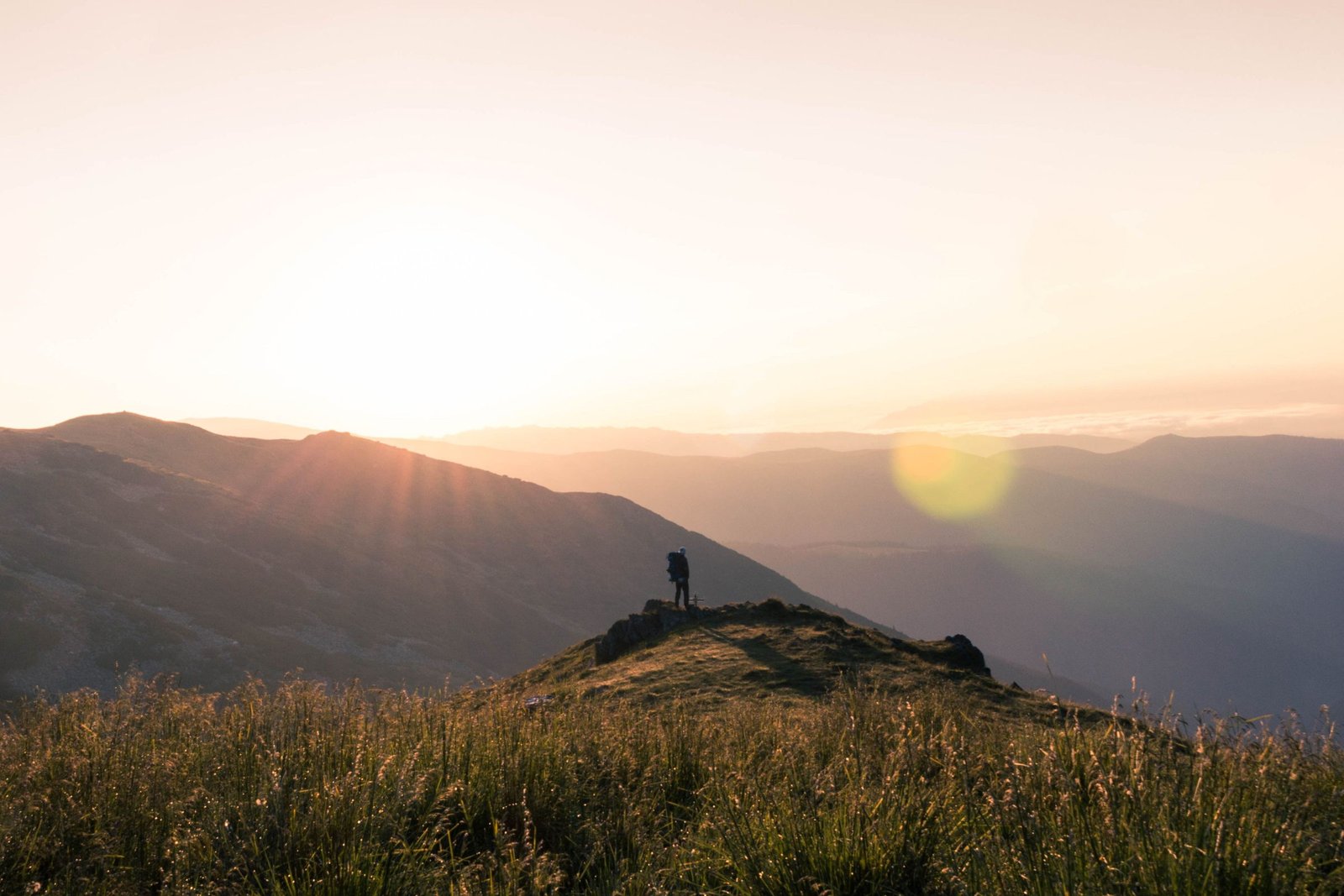 A lone traveller with a backpack seen from behind, standing on a ridge facing a range of mountains at sunrise