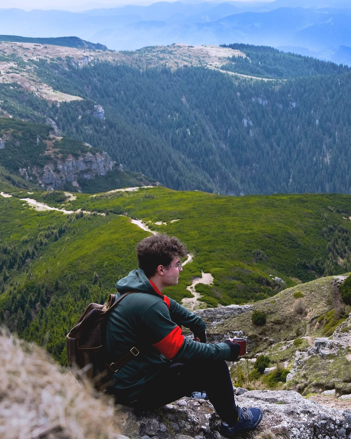 A traveller sitting on a rock ledge with a backpack beside them, looking out over a cloud-filled valley