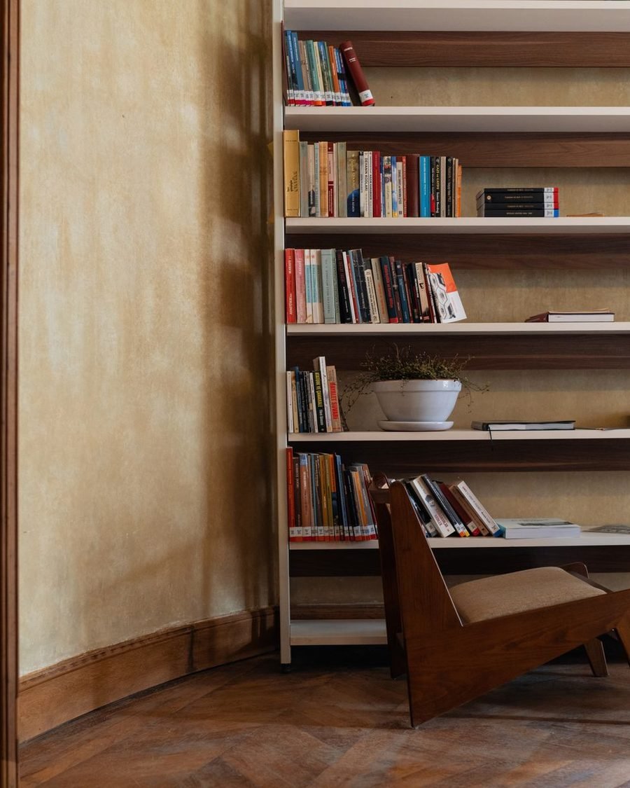 Reading room corner with full-height timber shelving, a low reading chair and warm daylight on a lime-plastered wall