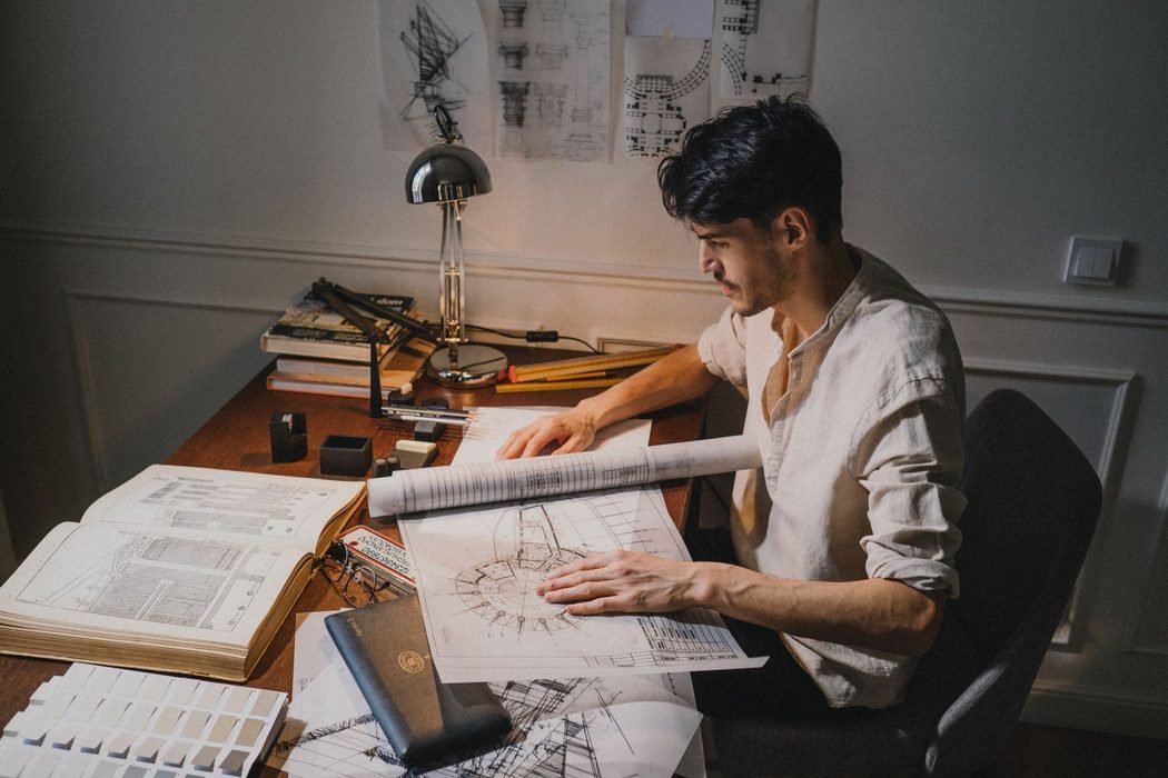 Architect at a desk under a lamp, unrolling a set of site drawings beside reference books
