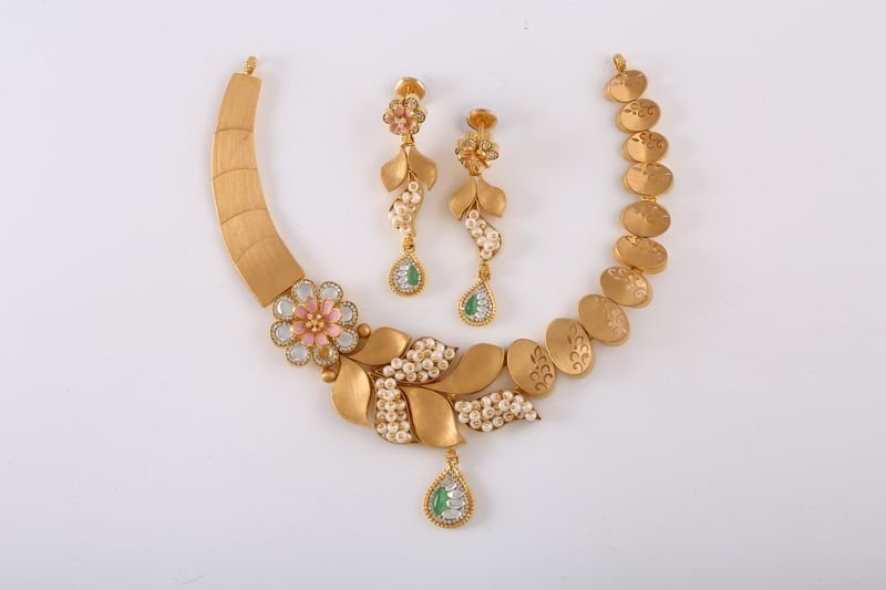 A matte-finish 22-karat gold necklace and earring set with pearl and green stone drops on a white surface