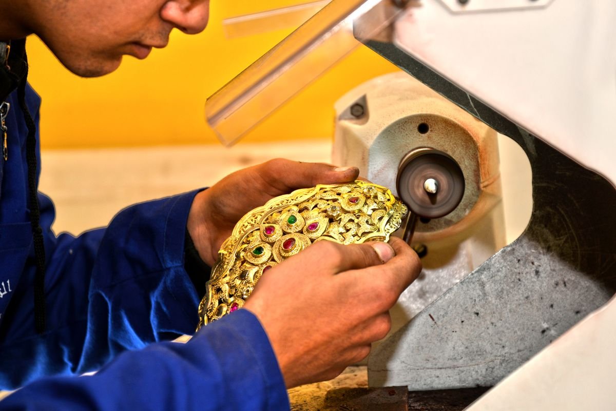 A goldsmith holding an ornate gold panel against a polishing wheel in the workshop