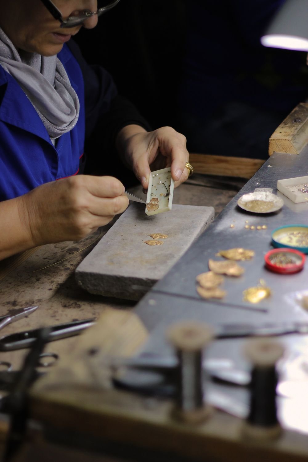 A jeweller setting small stones into a gold piece with tweezers at a workbench under a lamp