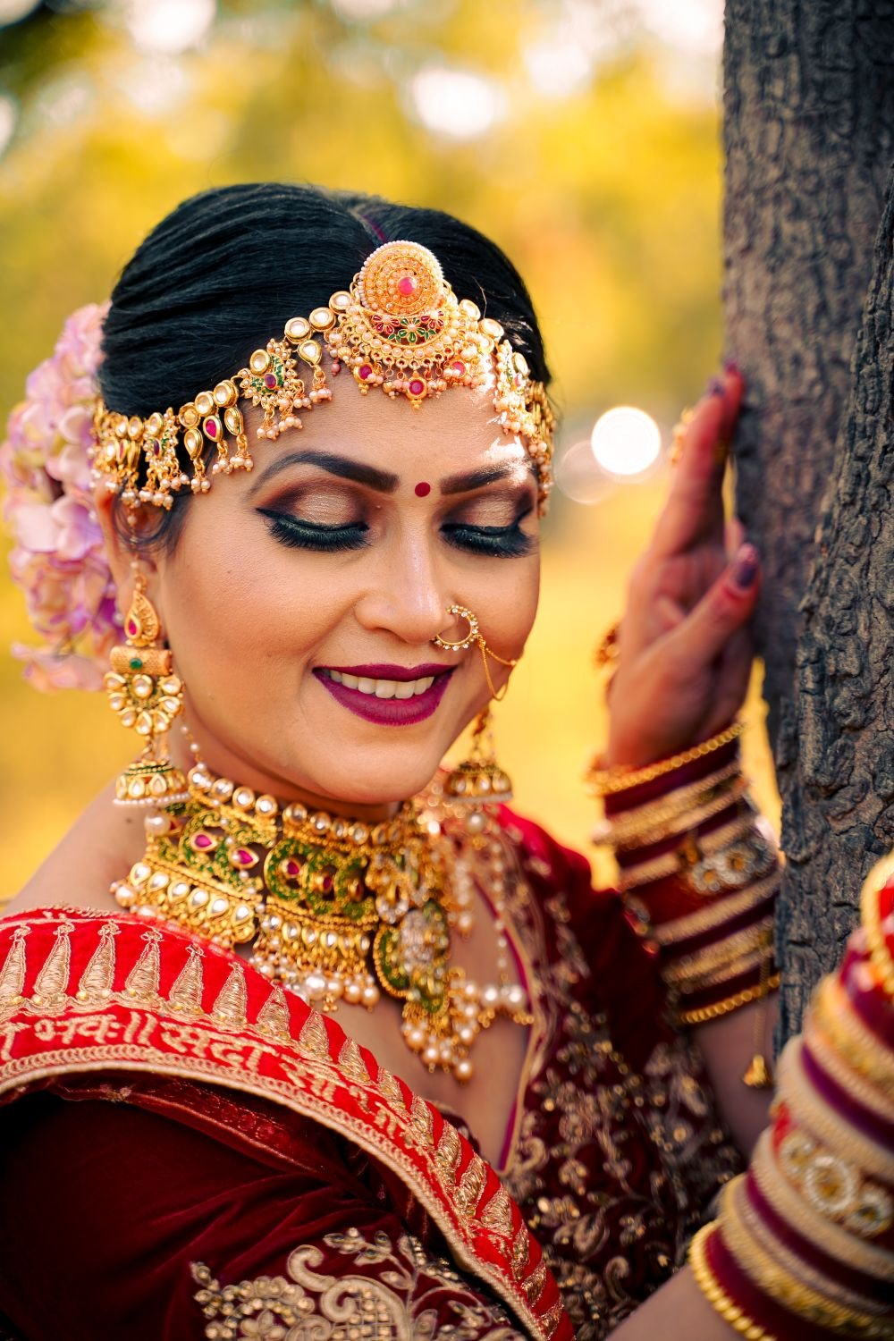 A bride in a red and gold outfit wearing a kundan matha patti, jhumkas and a choker, photographed outdoors