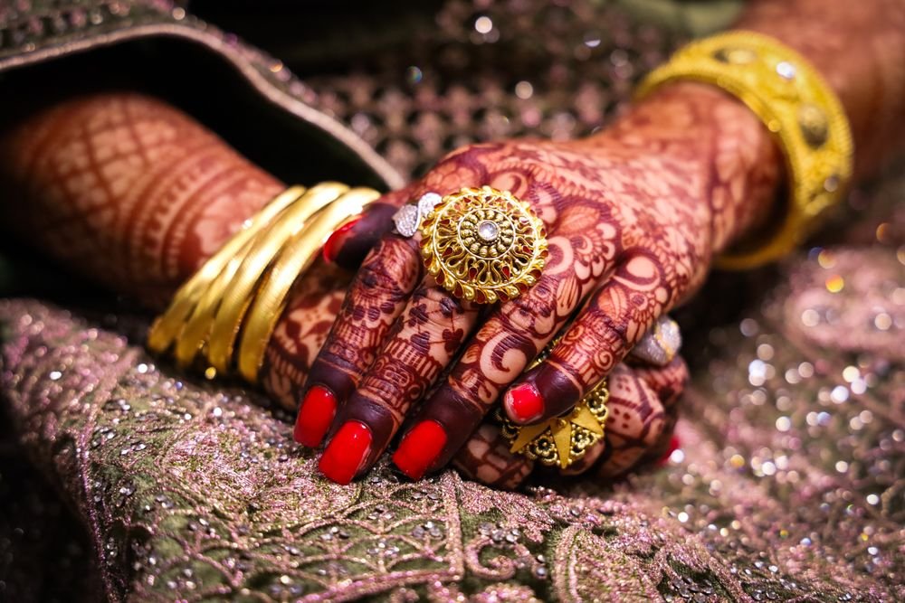 Hennaed hands resting on a green lehenga, wearing gold bangles and a large round cocktail ring