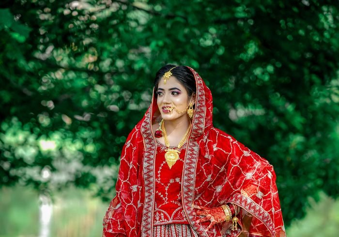 A bride in a red saree with gold bridal jewellery photographed against green foliage
