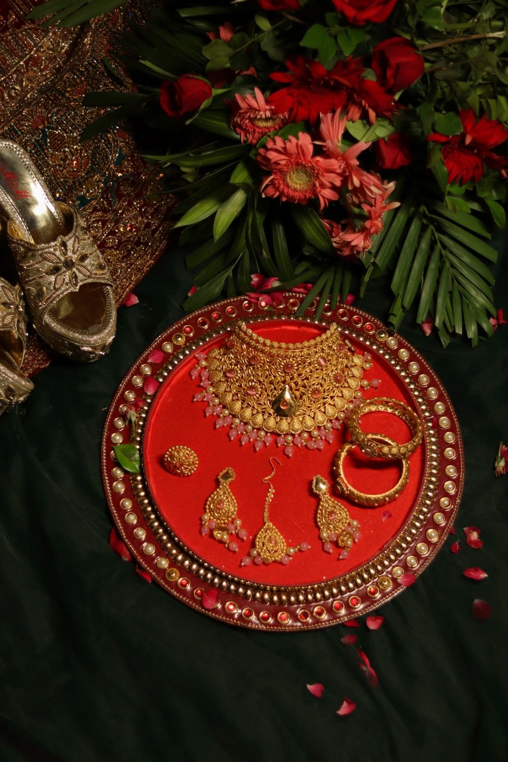 Gold bridal set — choker necklace, jhumka earrings, maang tikka and bangles — laid on red velvet thali with rose petals